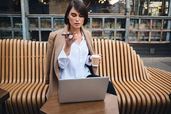 Молодая уверенная в себе стильная деловая женщина с ноутбуком записывает голосовое сообщение на мобильный телефон во время кофе-брейка в кафе на улице — стоковое фото