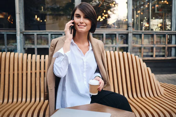 Молодая довольно стильная деловая женщина с ноутбуком счастливо разговаривает по мобильному телефону во время кофе-брейка в кафе на улице — стоковое фото