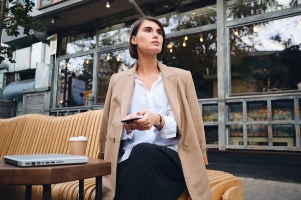 Молодая шикарная стильная деловая женщина с мобильным телефоном пристально смотрит в сторону во время кофе-брейка в кафе на улице — стоковое фото