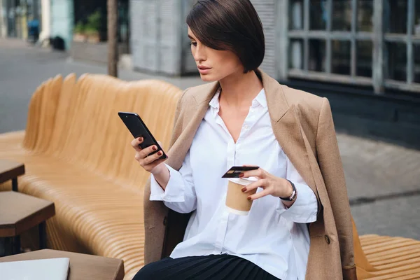 Ung, attraktiv forretningskvinne med kredittkort som bruker mobiltelefon under kaffepause på gaten – stockfoto