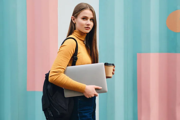 Bella ragazza studente casuale con computer portatile e caffè per andare pensieroso alla ricerca in macchina fotografica su sfondo colorato — Foto Stock