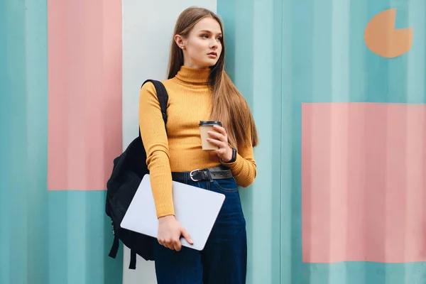 Attraktiv seriös student flicka med bärbar dator och kaffe för att gå tittar bort över färgglada bakgrund — Stockfoto