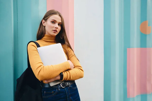 Attraente ragazza studente casuale con computer portatile pensieroso guardando in macchina fotografica su sfondo colorato — Foto Stock