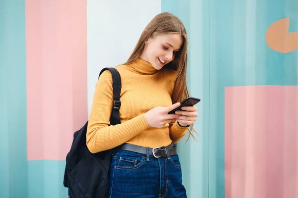 Atrakcyjny uśmiechnięty student dziewczyna radośnie za pomocą telefonu komórkowego na kolorowe tło zewnątrz — Zdjęcie stockowe