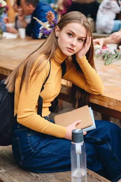 Menina estudante casual grave bonita com livro olhando atentamente na câmera ao ar livre — Fotografia de Stock