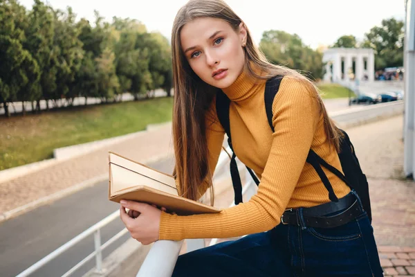 Menina estudante atraente com livro de texto pensativo olhando na câmera estudando no parque da cidade — Fotografia de Stock