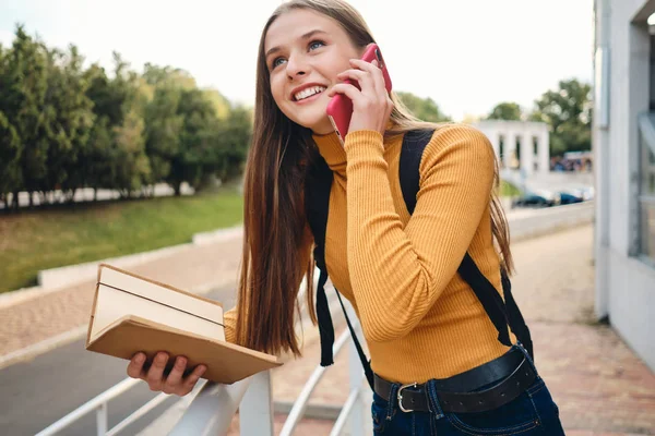 Atrakcyjny uśmiechnięta uczennica z notatnikiem szczęśliwie rozmawia przez komórkę studiuje w parku miejskim — Zdjęcie stockowe