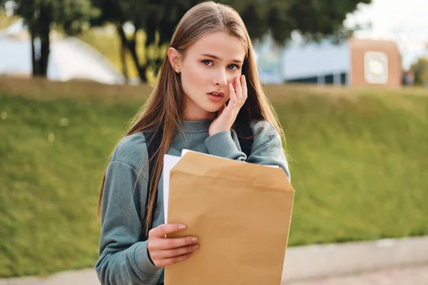 Portrait of upset casual student girl opening envelope with exams results sadly looking in camera in city park — Stock Photo, Image