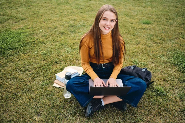 Bella sorridente ragazza studente casuale felicemente alla ricerca in macchina fotografica studiando con il computer portatile sul prato nel parco — Foto Stock