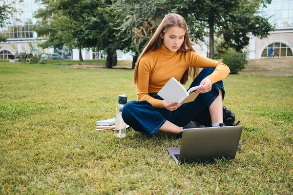 Vakker tilfeldig student leser bok med laptop på plen i parken – stockfoto