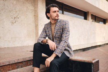 Zarif damalı ceketli, yakışıklı, ciddi bir adam. Düşünceli bir şekilde dışarı bakıyor.