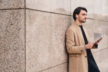 Dışarıda gazete okuyan, kablosuz kulaklıklı, bej ceketli yakışıklı bir adam.