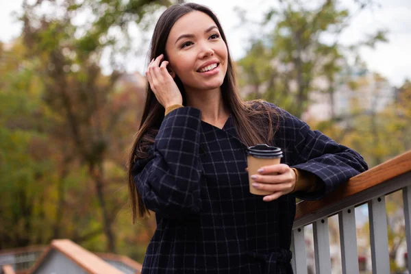 Молодая привлекательная женщина в пальто с кофе весело смотрит в парк — стоковое фото