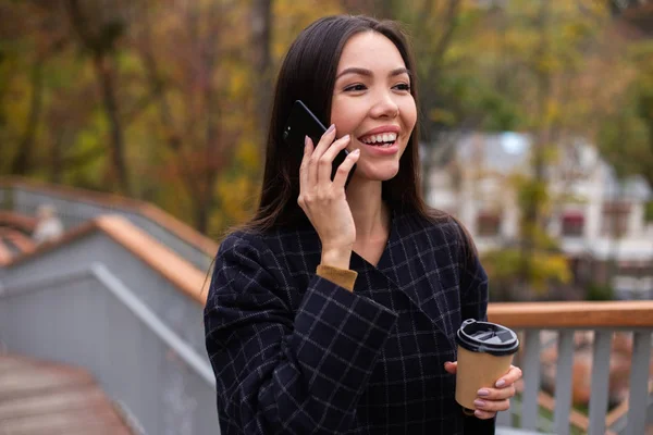 Ung, positiv, tilfeldig kvinne i frakk med kaffe som gledelig snakker på mobiltelefon i parken – stockfoto