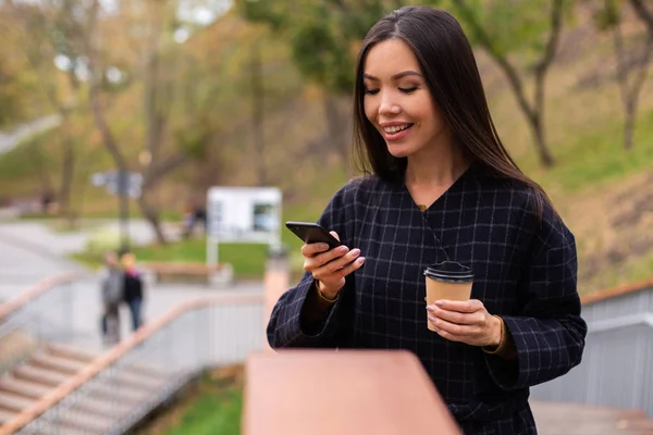 Молода красива випадкова жінка в пальто з кавою, щоб щасливо піти використовуючи мобільний телефон в міському парку — стокове фото