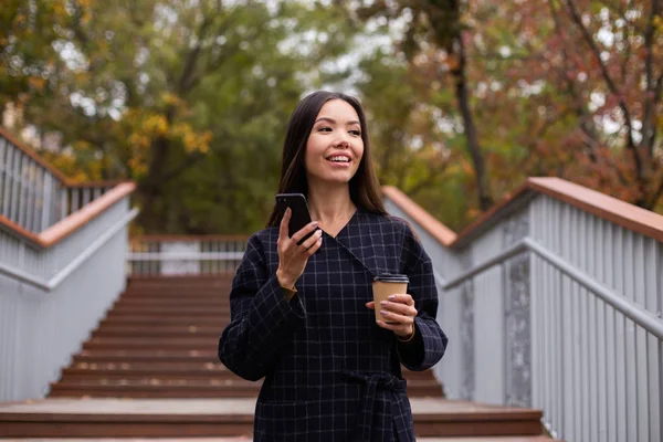 Joven mujer bastante casual en abrigo con café para llevar y teléfono celular mirando felizmente en el parque de la ciudad —  Fotos de Stock