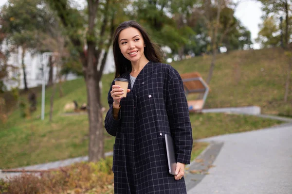 Молодая довольно случайная женщина в пальто с кофе, чтобы пойти и ноутбук счастливо глядя в парке — стоковое фото