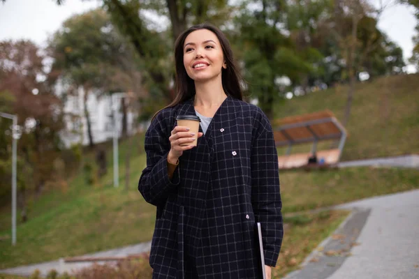 Ung, attraktiv kvinne i frakk med kaffe til å gå med og laptop lykkelig spaserer i byparken – stockfoto