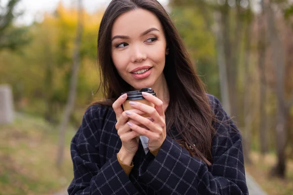 Retrato de mujer joven atractiva en abrigo con café para ir slyly mirando hacia otro lado en el parque de otoño — Foto de Stock