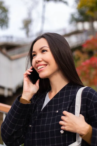 Вид сбоку на молодую улыбающуюся случайную женщину в пальто, счастливо разговаривающую по мобильному телефону в городском парке — стоковое фото