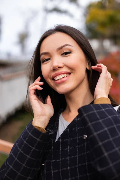 Портрет молодой красивой женщины в пальто, радостно смотрящей в камеру и разговаривающей по сотовому телефону в городском парке — стоковое фото