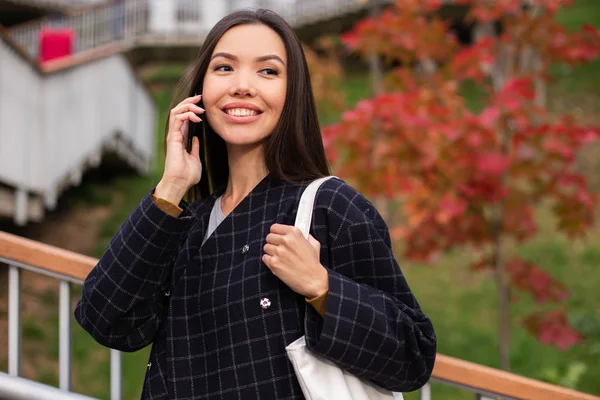 Молодая красивая случайная женщина в пальто радостно разговаривает по мобильному телефону в осеннем парке — стоковое фото