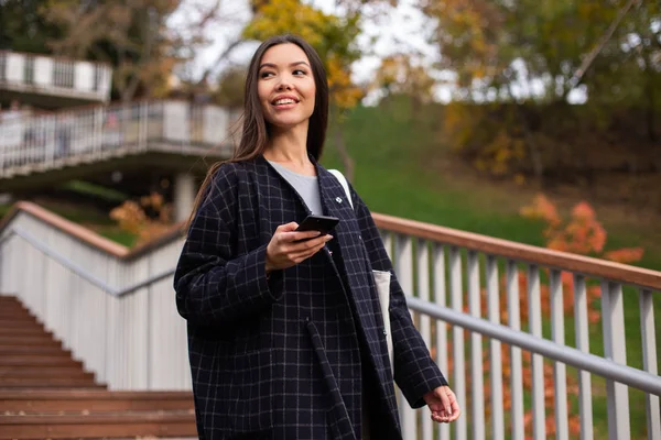 Mladý atraktivní usmívající se žena v kabátě s mobilem šťastně procházky v městském parku — Stock fotografie