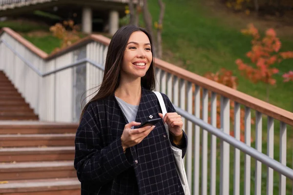 Ung, munter kvinne i frakk med mobiltelefon som går lykkelig i byparken – stockfoto