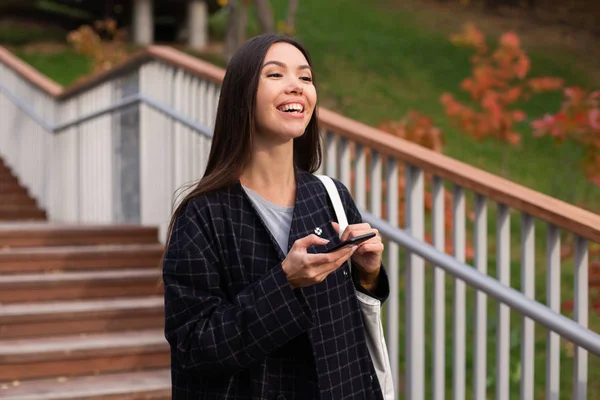 Wanita muda yang santai dengan mantel dengan ponsel dengan senang hati berjalan di taman musim gugur — Stok Foto