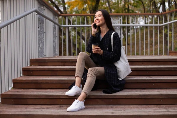 Молодая привлекательная случайная женщина с кофе идти радостно говорить по сотовому телефону сидя на лестнице в парке — стоковое фото
