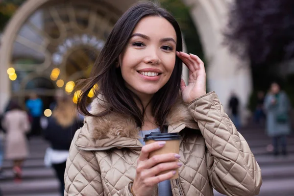 Muda cukup tersenyum santai wanita di bawah jaket dengan kopi untuk pergi bahagia mencari di kamera di jalan kota — Stok Foto