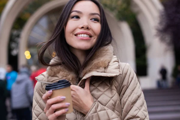 Ung, vakker, avslappet kvinne i dunjakke med kaffe som gledelig ser i kamera på byens gate – stockfoto