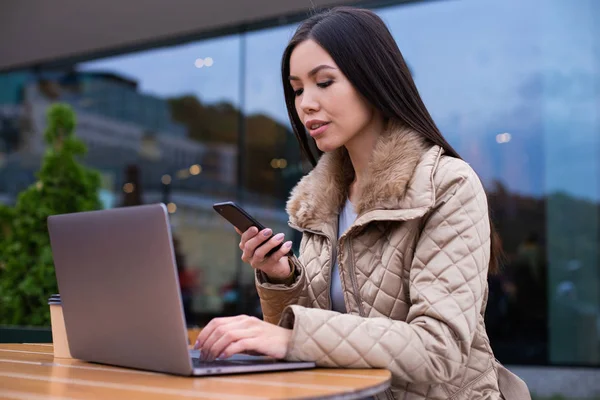 Wanita muda yang percaya diri dengan jaket yang bekerja pada laptop dengan ponsel di jalan di kafe — Stok Foto