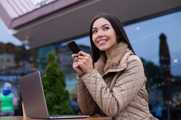 Young attractive woman in down jacket slyly looking away using cellphone in cafe on street — Stock Photo, Image