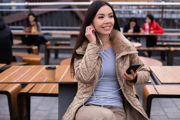 Wanita muda yang santai ceria di bawah jaket senang mendengarkan musik di earphone di luar ruangan ponsel — Stok Foto