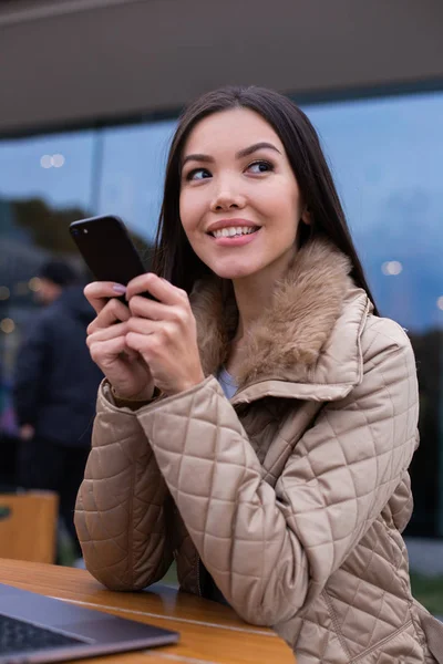 En ung flørtete kvinne i jakke som ser seg bort med mobiltelefon på gata – stockfoto