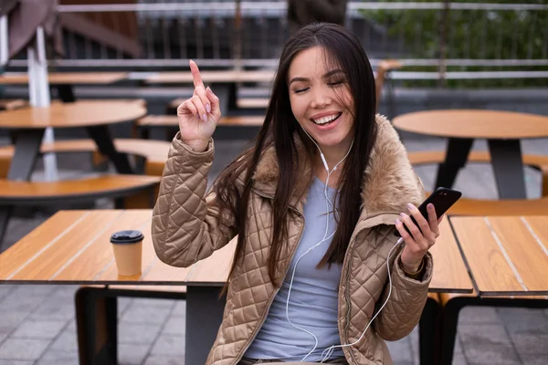 Wanita muda yang santai ceria mengenakan jaket dengan senang hati menari mendengarkan musik di earphone di luar ruangan ponsel — Stok Foto