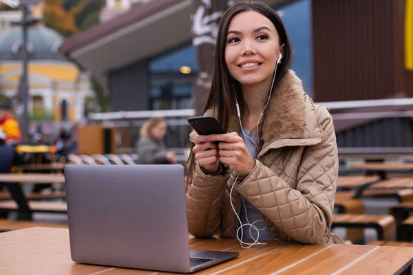 Wanita muda yang kasual dengan laptop dengan senang hati menggunakan ponsel di kafe di jalan — Stok Foto