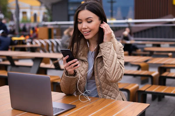 Ung, munter kvinne i dunjakke, lyttende musikk i øretelefoner på mobil i kafe på gaten – stockfoto