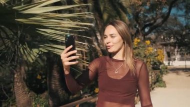 Şehir parkında yürürken ailesiyle görüntülü konuşma yapmak için akıllı telefonu kullanan çekici bir kız. Mavi gözlü güzel sarışın kız video mesajı kaydediyor.