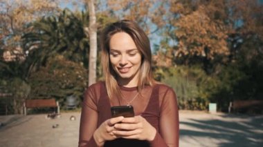 Güzel bir kızın parkta yürürken arkadaşlarıyla mesajlaşmak için akıllı telefon kullanmasının görüntüsü.