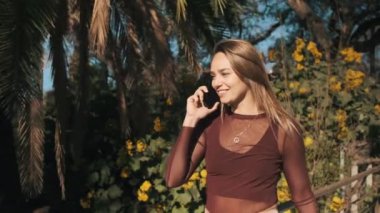 Çekici, gülümseyen bir kızın güzel bir parkta palmiyelerle yürürken ailesiyle telefonda mutlu bir şekilde konuşmasının görüntüsü. Güzel sarışın kız telefonda bir arkadaşıyla sohbet ediyor.
