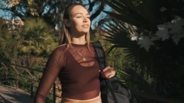 Futbol ve sırt çantalı çekici bir kızın idmandan sonra şehir parkında yürüdüğü orta boy bir fotoğraf. Güzel, çekici, sarışın kız kameraya bakıp güneşin tadını çıkarıyor.