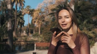Parkta yürürken akıllı telefondan mutlu bir şekilde sesli mesaj kaydeden çekici bir kızın çekimini izliyorum. Açık havada sesli haberci kullanarak gülümseyen mavi gözlü güzel sarışın kız.