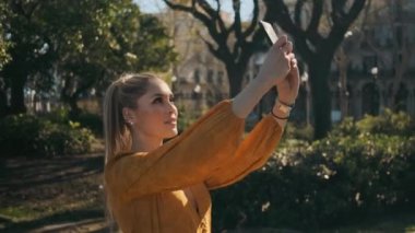 Şehir parkında yürürken akıllı telefondan fotoğraf çeken güzel bir kadının fotoğrafını çekiyorum. Çekici bayan dışarıda selfie çekiyor.