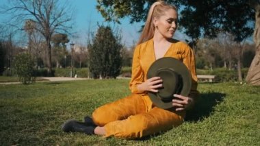 Turuncu tulumlu, şapkalı, şehir parkındaki çimlerde dinlenen genç ve güzel bir kadın.