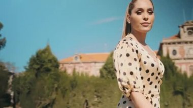 Etrafta polka noktalı elbise giymiş genç ve şık bir kadının, parkta yürürken kameraya güvenle baktığı bir fotoğraf var.
