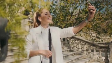 Beyaz smokinli, şık, genç bir kadın şehrin eski parkındaki merdivenlerde akıllı telefondan mutlu bir şekilde selfie çekiyor.
