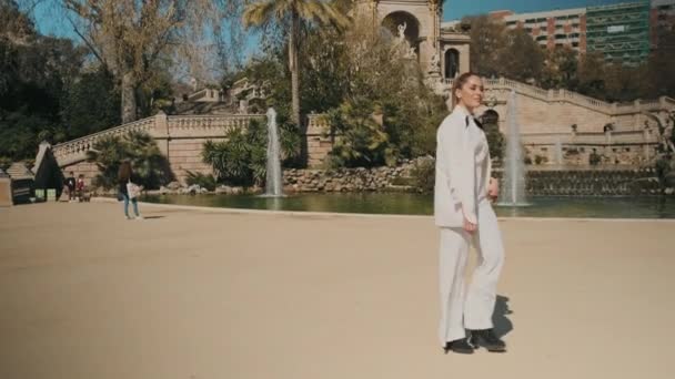 白いスーツの若い豪華なエレガントな女性は自信を持って古典的な建築物の上に古い美しい都市公園に沿って歩く — ストック動画