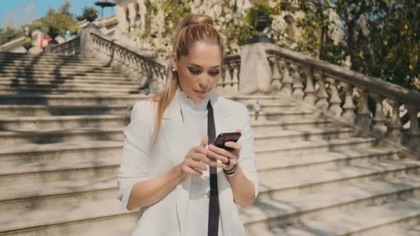 位于旧城公园的楼梯上 年轻而优雅的女子 穿着经典套间 自信地站在楼梯间的智能手机上自拍 — 图库视频影像
